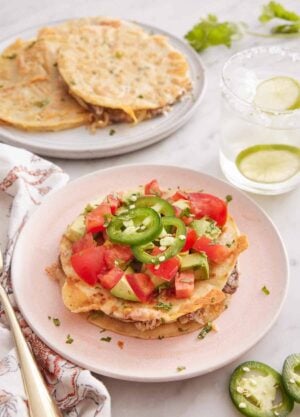 A plate of mulita topped with tomatoes, avocados, and jalapenos. A drink in the back and more mulitas on a plate.