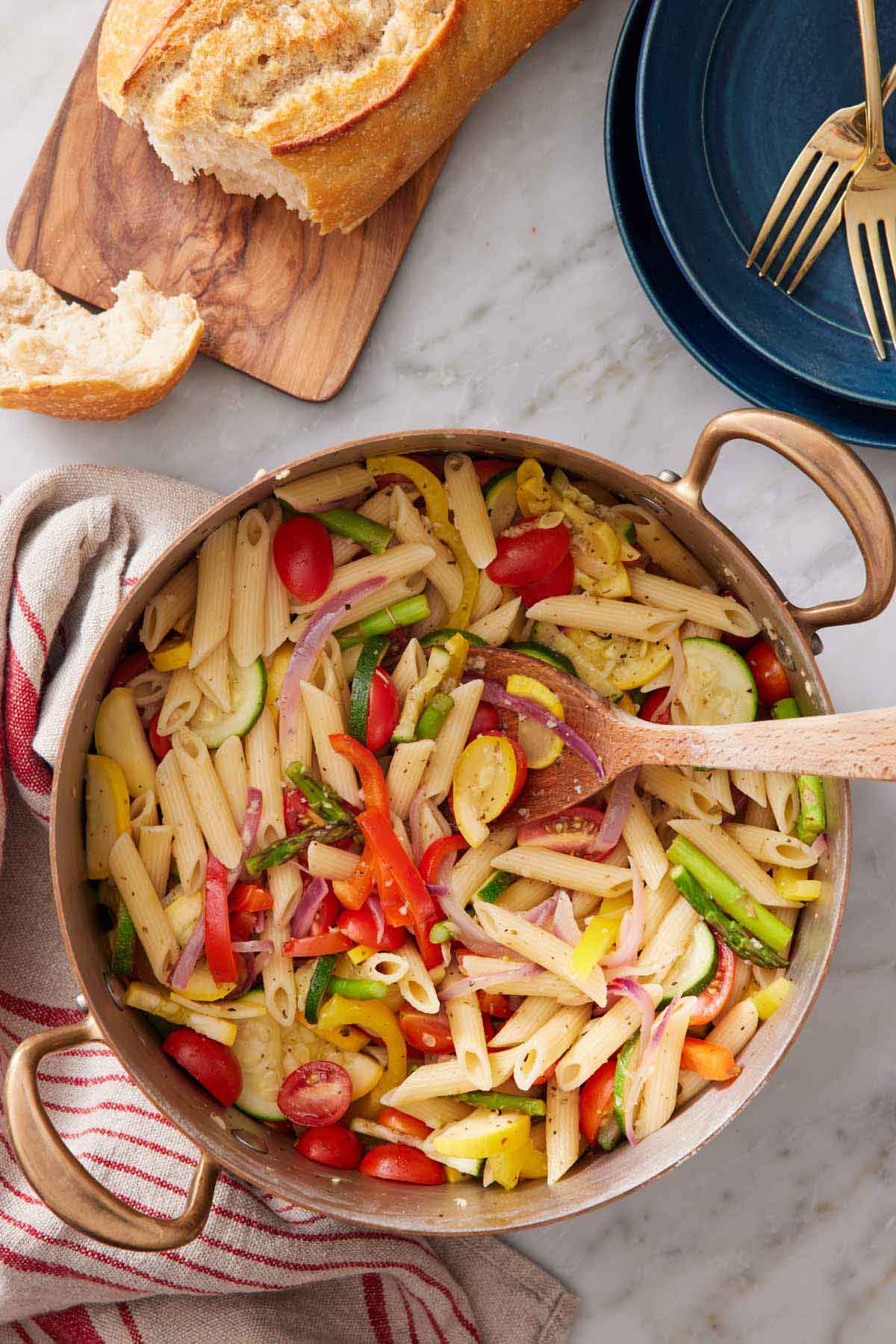 Overhead view of a pot of pasta primavera with some torn bread and a stack of plate with forks on the side.
