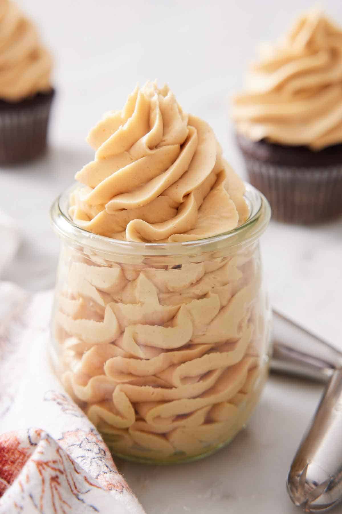 A glass jar with peanut butter frosting piped inside. Two more cupcakes with frosting on top.