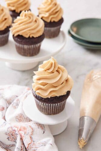 A cupcake with peanut butter frosting on top with more frosted cupcakes in the background. A piping bag and plates off to the side.