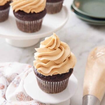 A cupcake with peanut butter frosting on top with more frosted cupcakes in the background. A piping bag and plates off to the side.