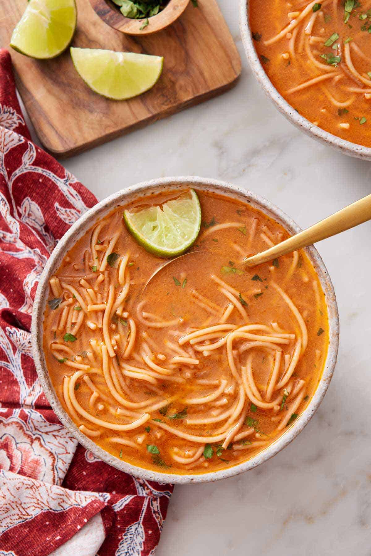 Overhead view of a bowl of sopa de fideo with a spoon and lime inside. Some sliced lime and another bowl of soup on the side.