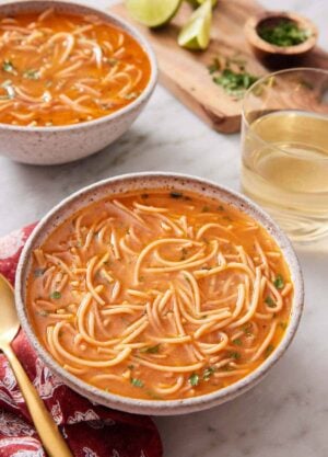 A bowl of sopa de fideo with a drink and second bowl in the background with some garnish on the side.