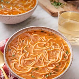 Pinterest graphic of a bowl of sopa de fideo with a drink and second bowl in the background with some garnish on the side.