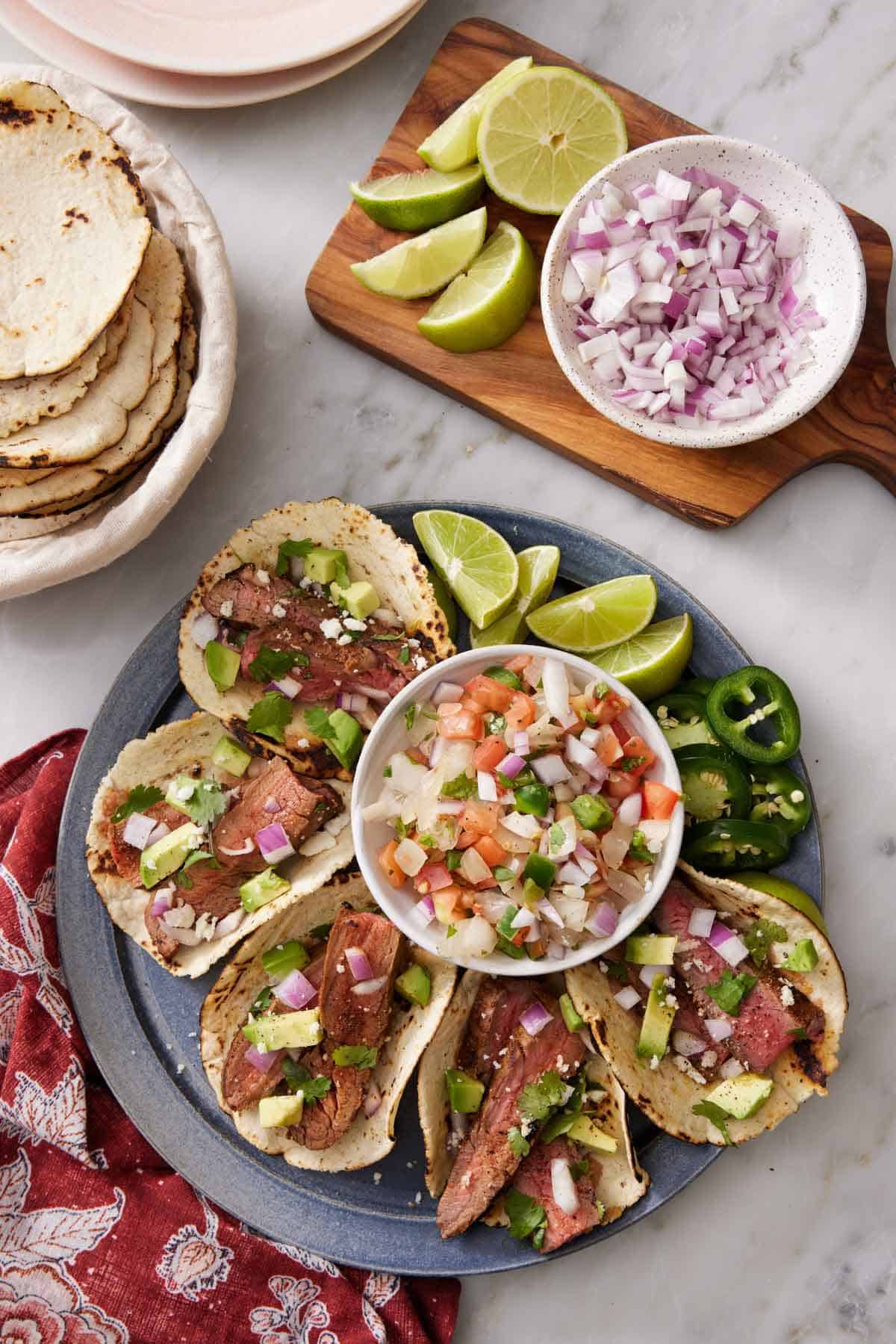 Overhead view of a platter of steak tacos along with a bowl of salsa, lime wedges, and jalapeno slices. A board with more toppings and bowl of tortilla to the side.