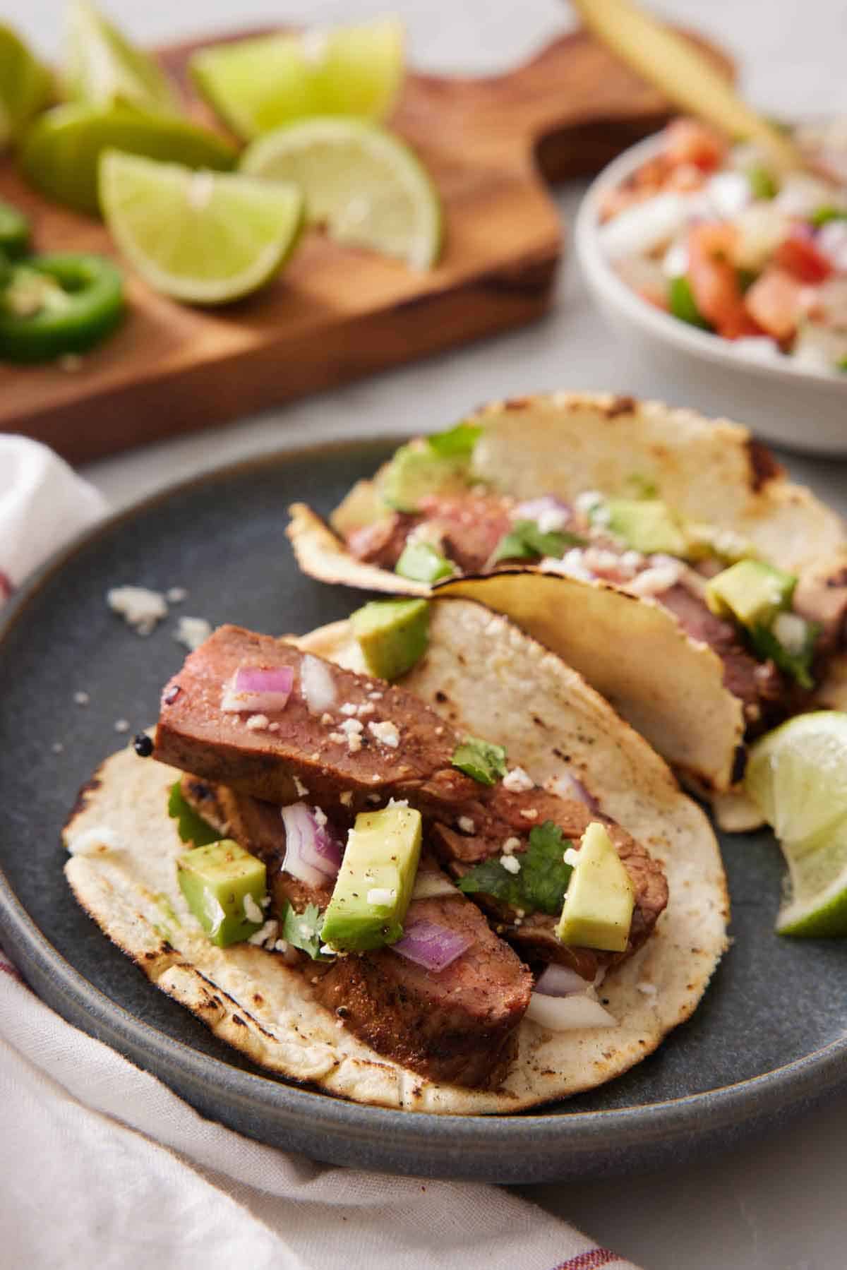 A close up view of a steak taco on a plate with a second one behind it. A bowl of salsa and cut limes in the background.