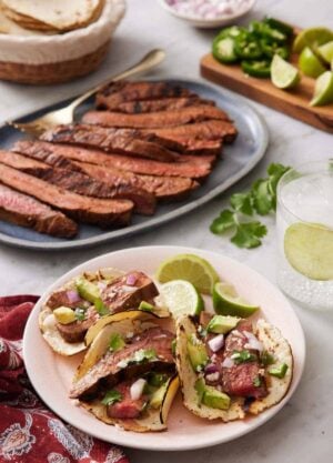 A plate with three steak tacos with lime wedges with a platter of sliced steak, bowl of tortilla, and chopped garnish in the background.