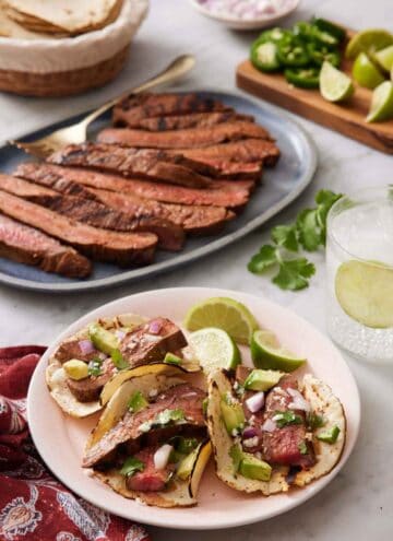 A plate with three steak tacos with lime wedges with a platter of sliced steak, bowl of tortilla, and chopped garnish in the background.