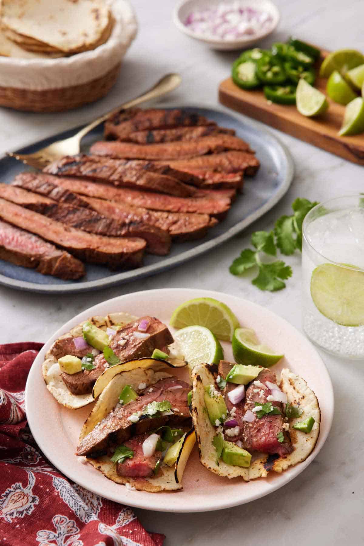 A plate with three steak tacos with lime wedges with a platter of sliced steak, bowl of tortilla, and chopped garnish in the background.