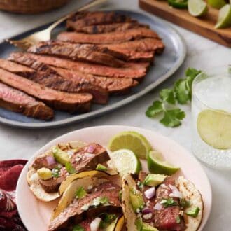 Pinterest graphic of a plate with three steak tacos with lime wedges with a platter of sliced steak in the back along with a drink and garnishes.