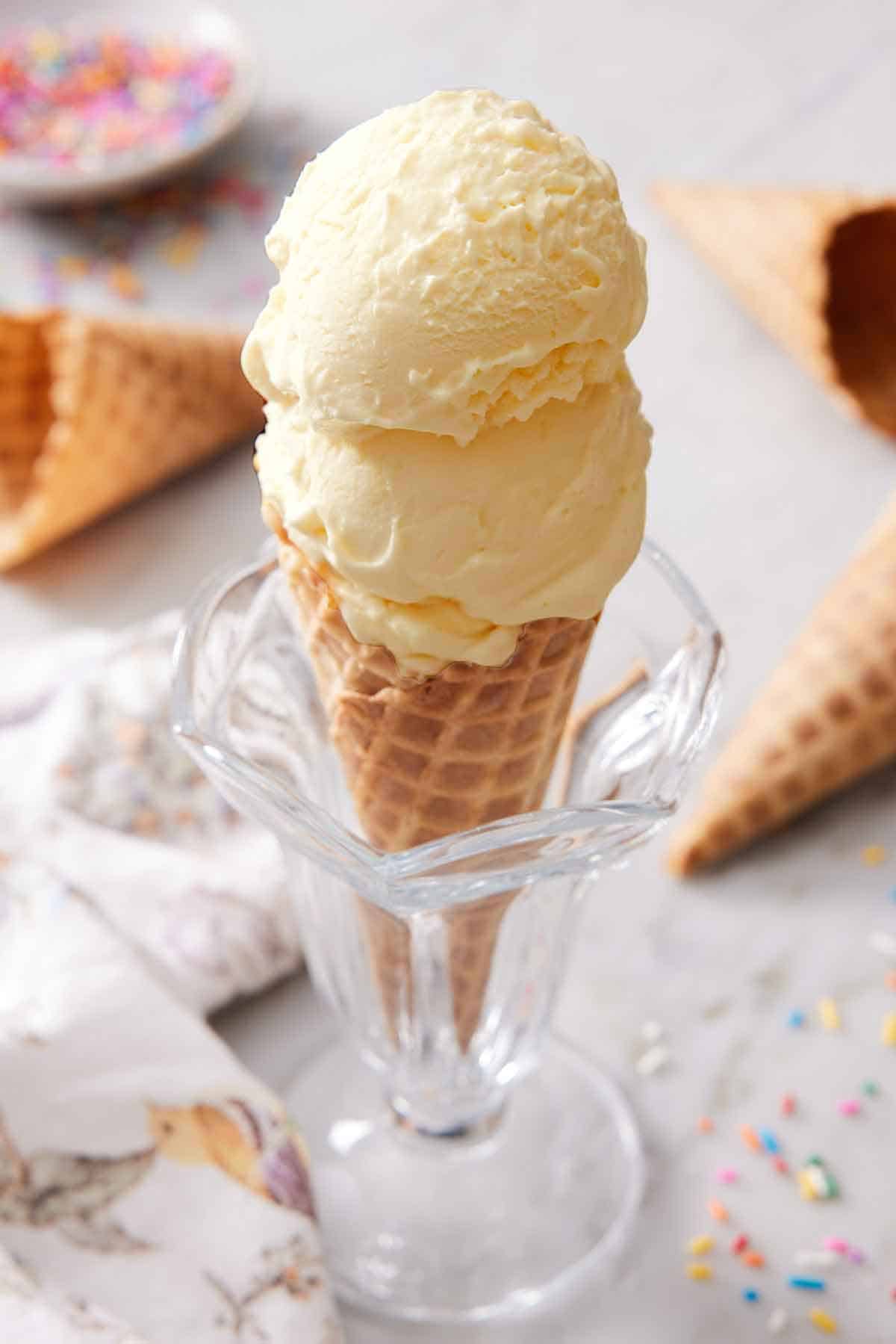 An ice cream cone with two scoops of vanilla ice cream. Cones and sprinkles scattered around.