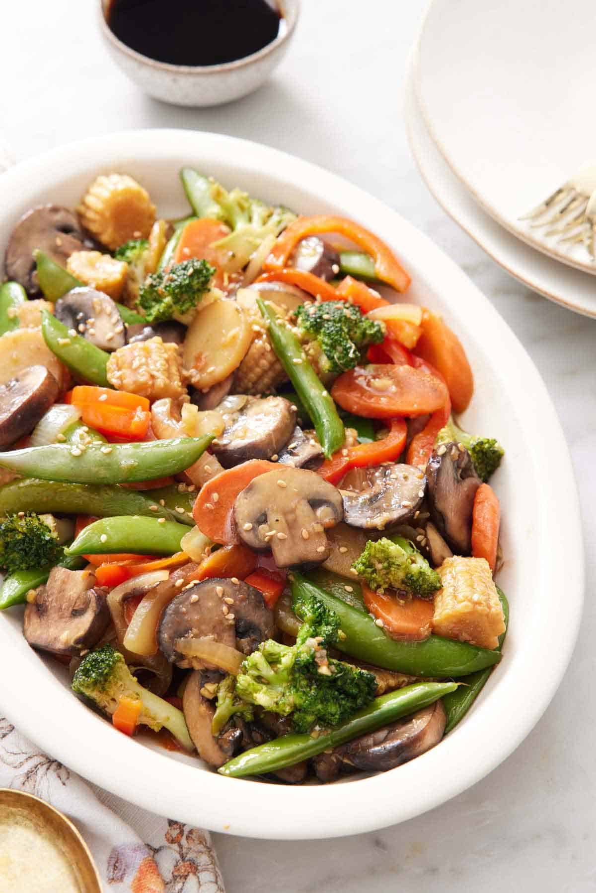 A platter of vegetable stir fry with a bowl of sauce in the back.