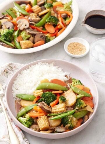 A plate of vegetable stir fry with rice. A platter with more vegetable stir fry in the background along with a bowl of sesame seeds and sauce.