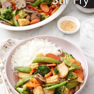 Pinterest graphic of a plate of vegetable stir fry with rice. A platter with more vegetable stir fry in the background. A bowl of sesame seeds and sauce in the background.