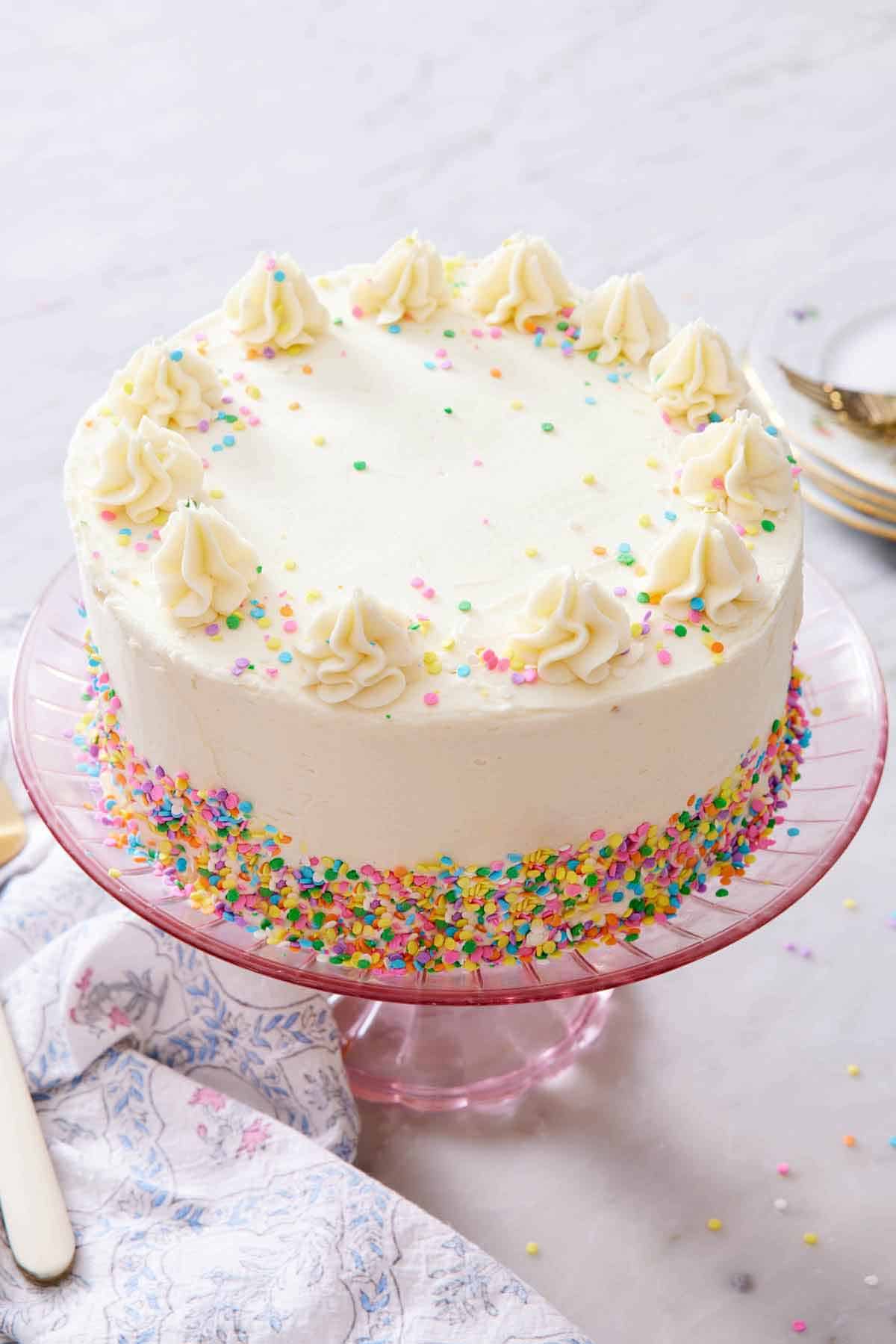 A white cake on a pink cake stand with dollops of frosting on the edge and rainbow sprinkles scattered around and on the edge.