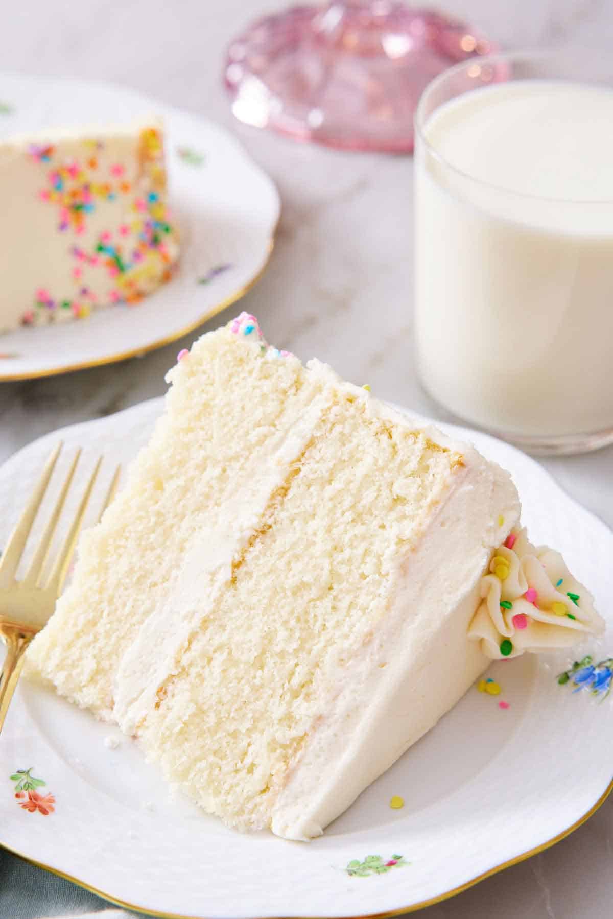A slice of white cake lying on its side on a plate, showing the two layers with frosting in between. Glass of milk in the background.