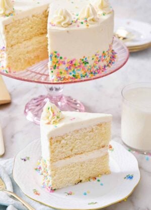 A slice of white cake on a plate with the rest of the cake in the background on a cake stand.