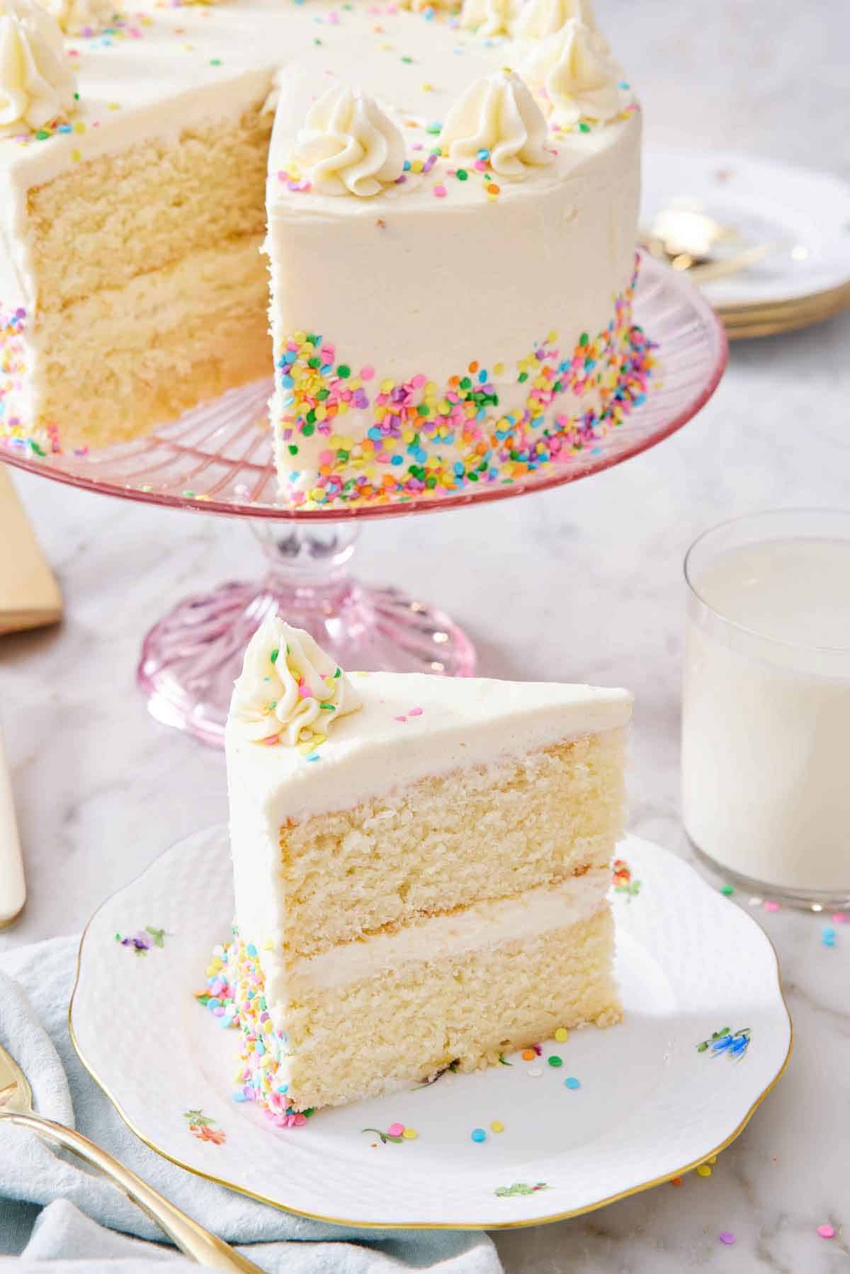 A slice of white cake on a plate with the rest of the cake in the background on a cake stand.