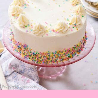 Pinterest graphic of a white cake on a pink cake stand with dollops of frosting on top and rainbow sprinkles scattered around and on the edge.