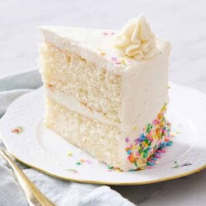 A slice of white cake on a plate with a dollop of frosting on top and rainbow sprinkles.