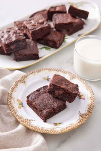 A plate with two pieces of air fryer brownies with a glass on milk in the back along with a platter more.