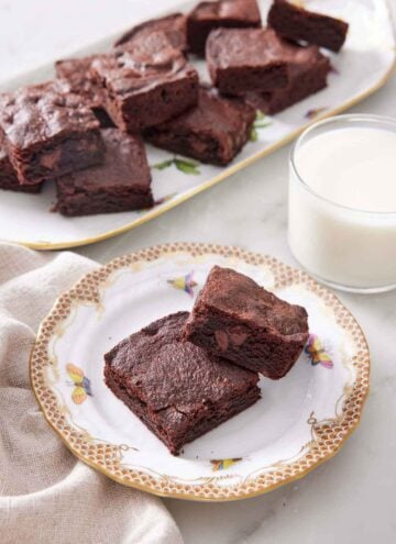 A plate with two pieces of air fryer brownies with a glass on milk in the back along with a platter more.