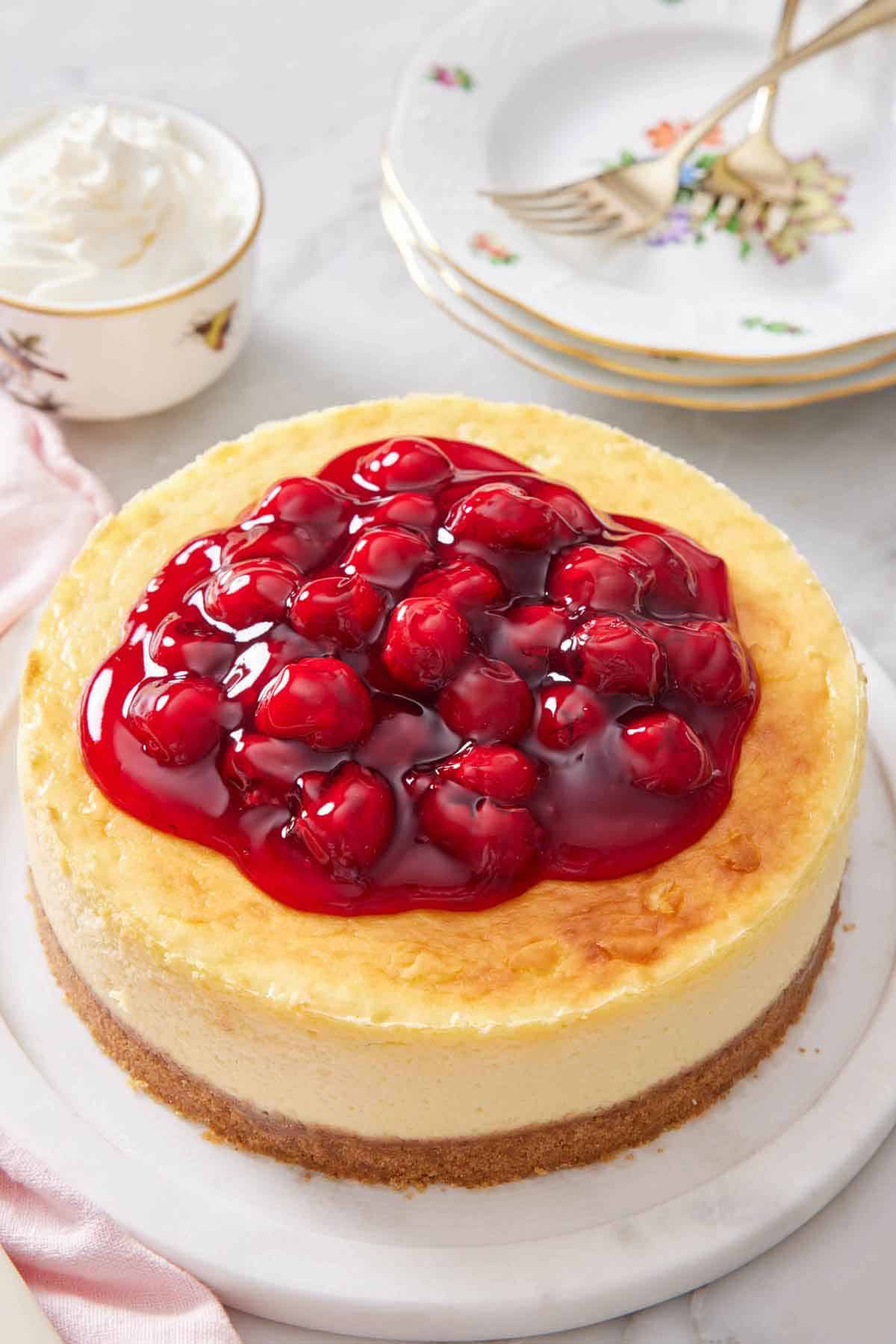 An air fryer cheesecake with cherry sauce on top. Stack of plates and forks in the background along with some whipped cream.