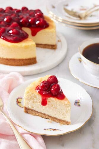 A slice of air fryer cheesecake on a plate with cherry sauce on top. The rest of the cheesecake in the background along with a mug of coffee.