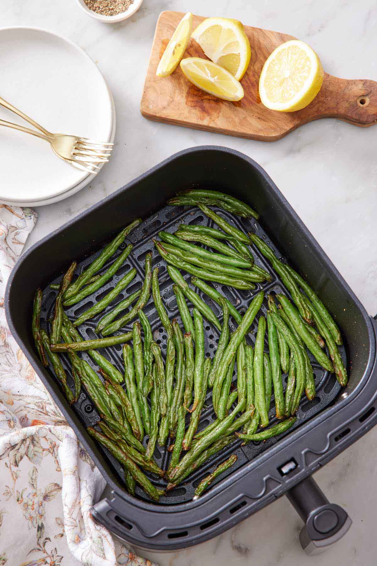 An overhead photo of green beans in an air fryer basket. A cut lemon off to the side along with a stack of plates and forks.