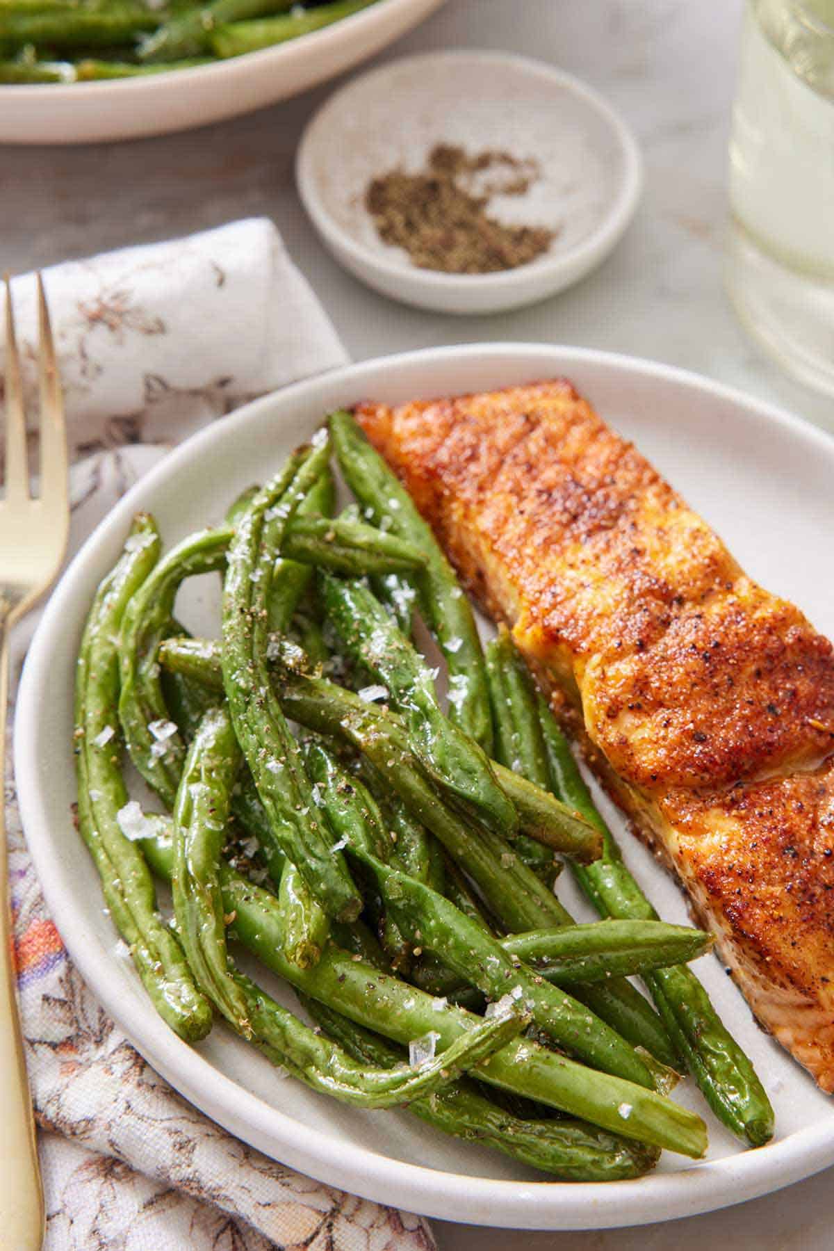 A plate with air fryer green beans with salmon. A small bowl of pepper and a drink in the background.