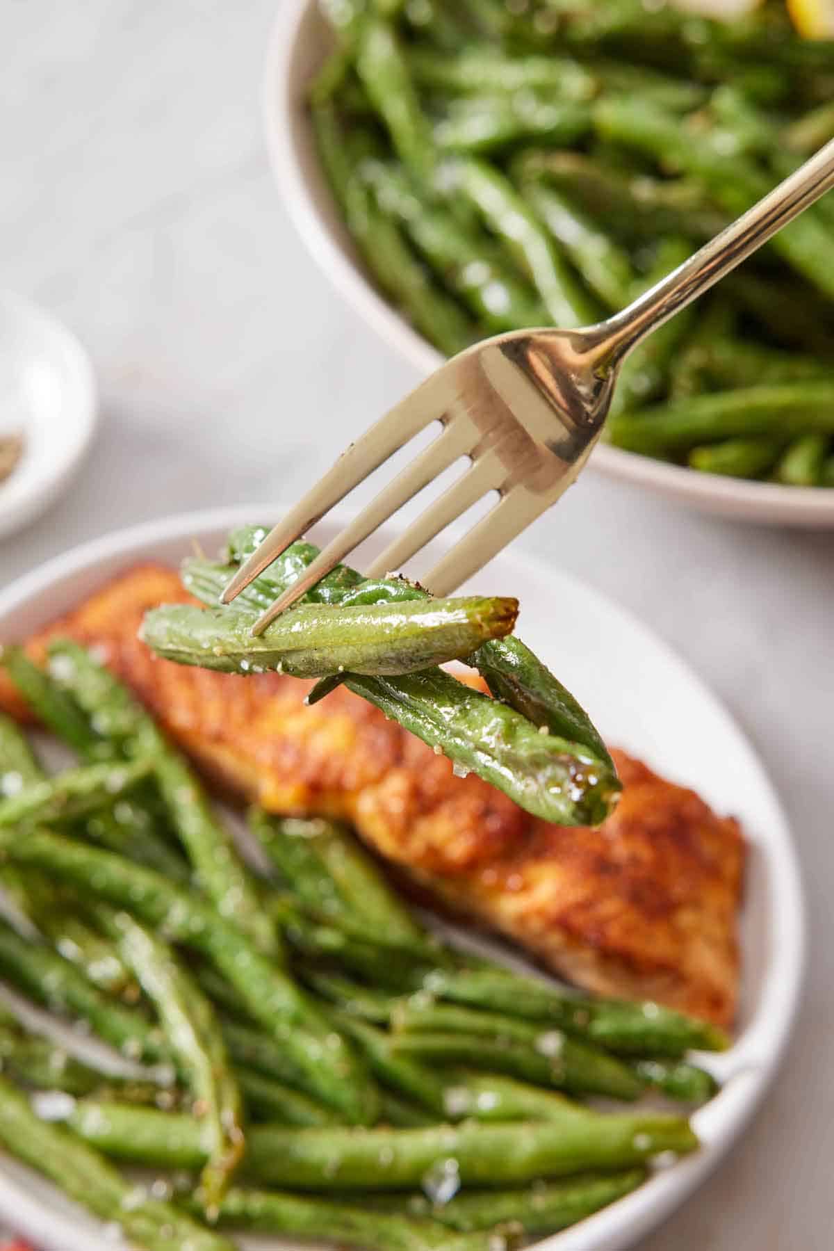A fork lifting up a three pieces of air fryer green beans from a plate with green beans and salmon.