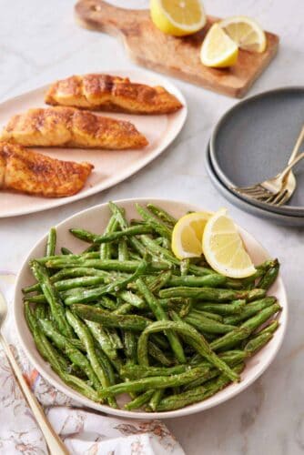 A plate of air fryer green beans with two lemon wedges. Platter of salmon in the back along with cut lemons and plates and forks.