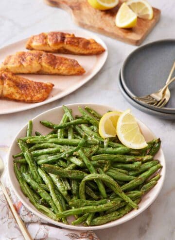 A plate of air fryer green beans with two lemon wedges. Platter of salmon in the back along with cut lemons and plates and forks.