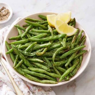 A plate of air fryer green beans with lemon wedges along with a fork on the side and bowl of pepper.