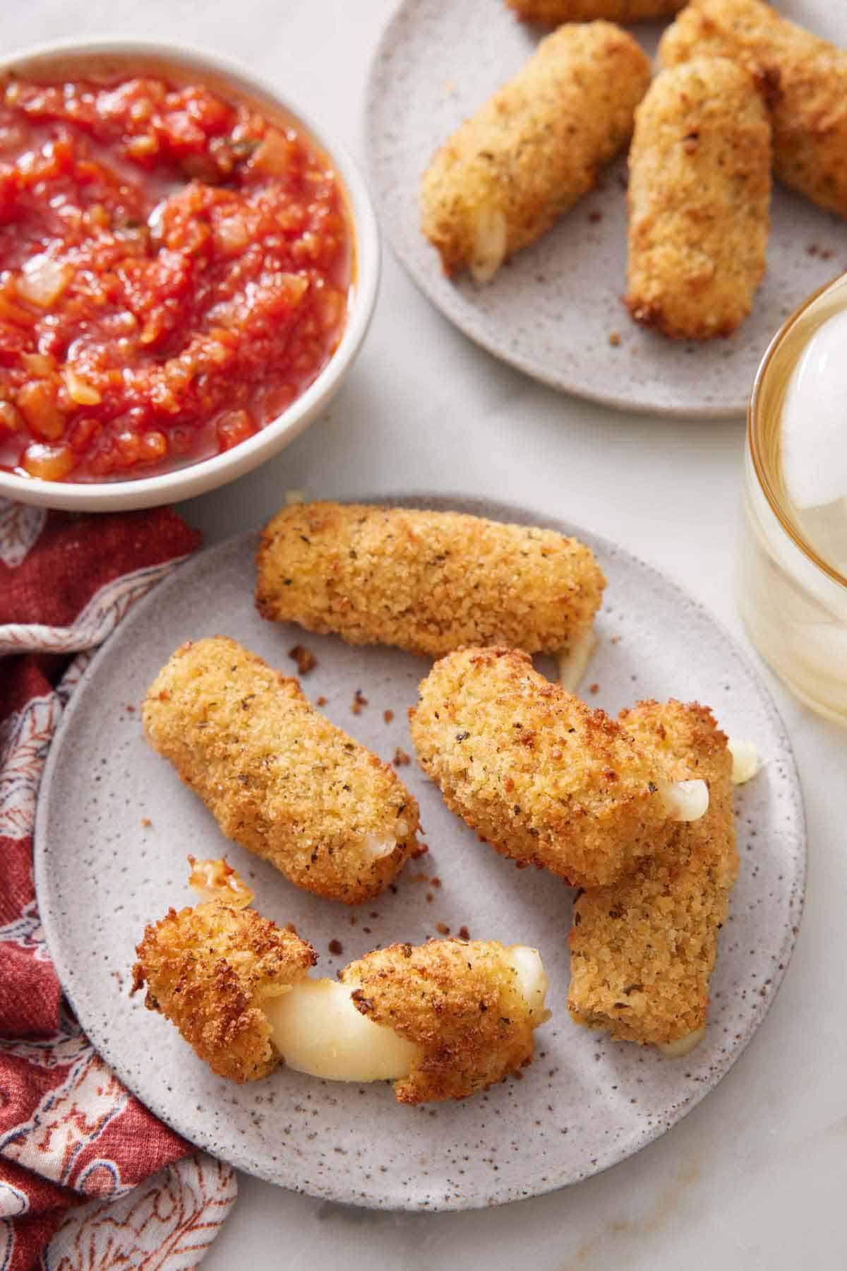 A plate of air fryer mozzarella sticks with one torn in half, showing the cheese stretched. A bowl of marinara sauce and more cheese sticks in the background.