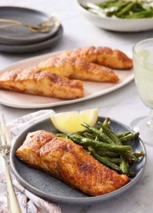 A plate with air fryer salmon along with green beans and a lemon wedge. A platter of more salmon in the background and plate of green beans.