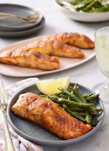 A plate with air fryer salmon along with green beans and a lemon wedge. A platter of more salmon in the background and plate of green beans.
