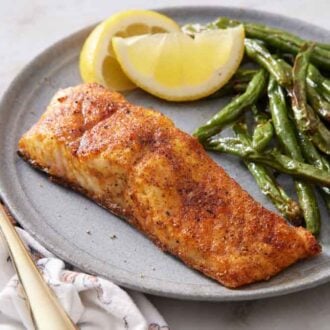 A plate with a piece of air fryer salmon with green beans and lemon wedges.