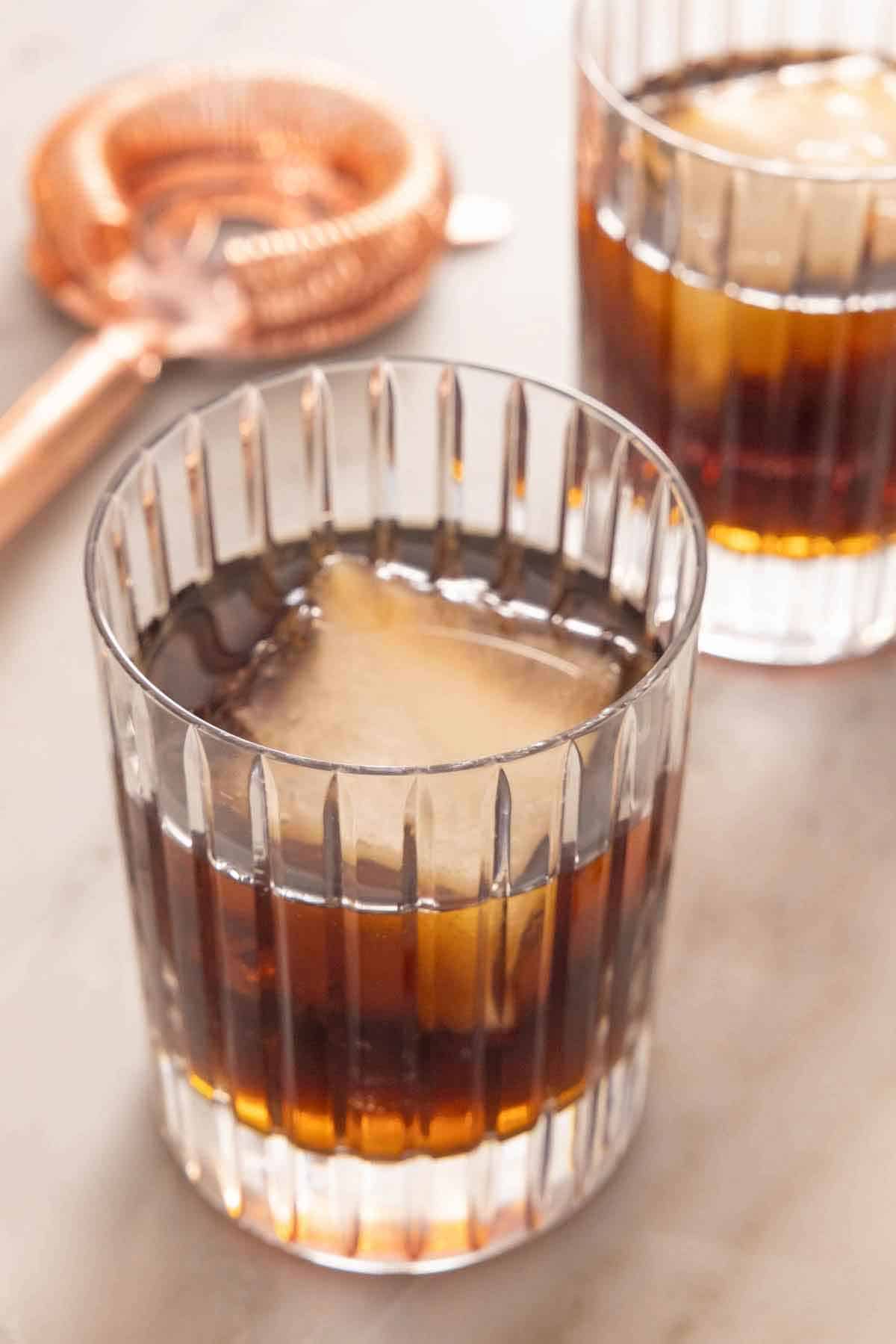 A glass of Black Russian with one square ice cube. A second one in the background along with a cocktail strainer.