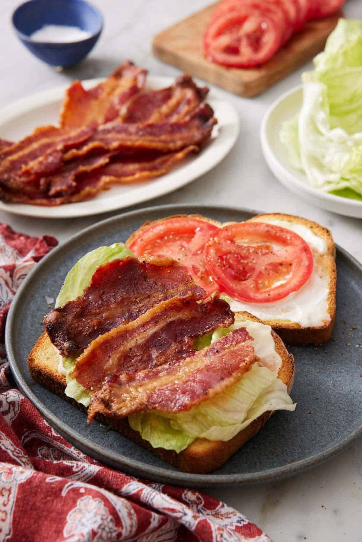A plate with two slices of bread with mayo, sliced tomatoes, lettuce, and candied bacon. More candied bacon in the background along with lettuce and tomatoes.