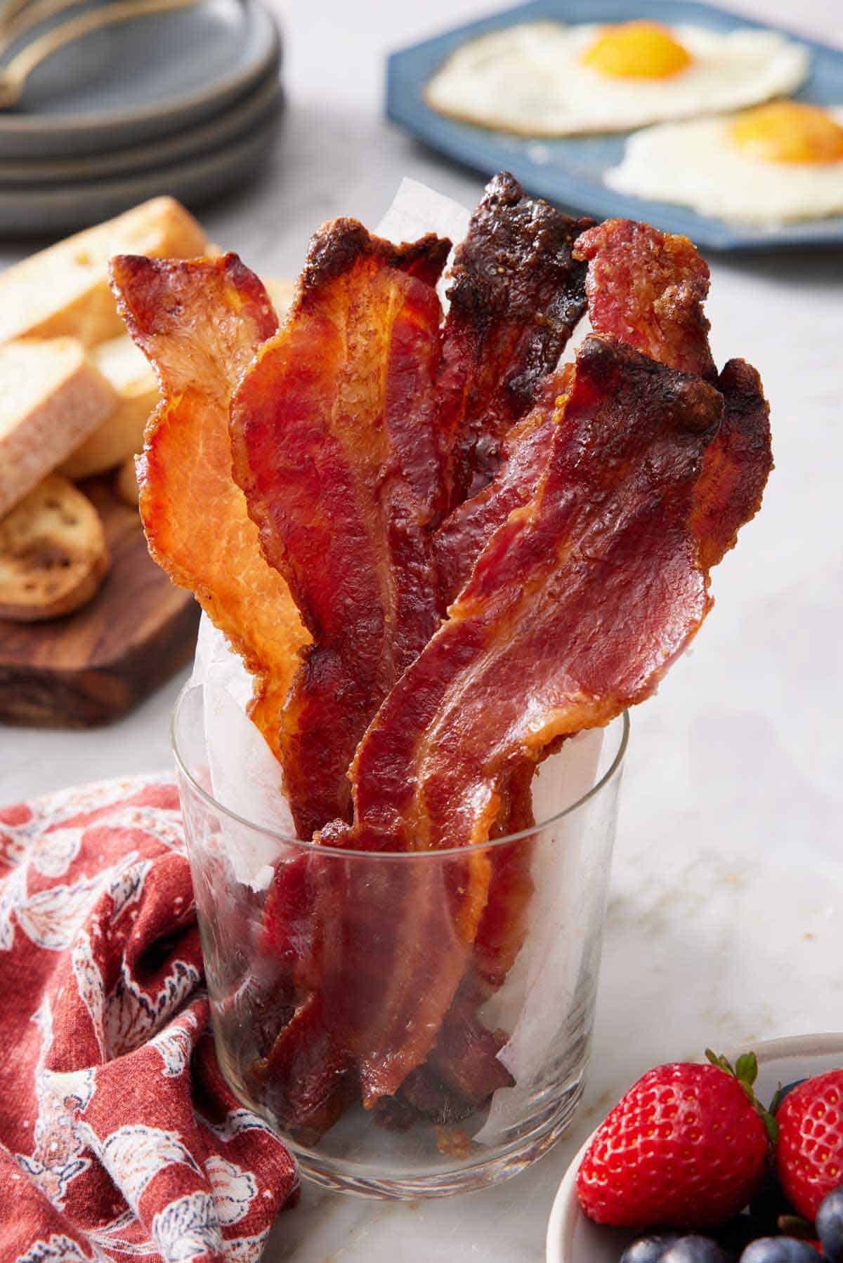 A glass cup of candied bacon. A platter in the background with fried eggs, sliced bread, and a stack of plates.