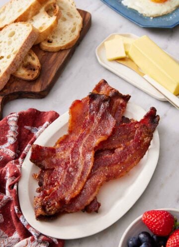 A plate of candied bacon with butter, bread, berries, and eggs around it.