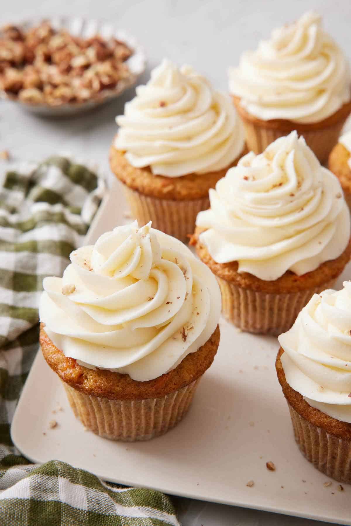 Multiple carrot cake cupcakes on a square platter with a checkered napkin beside them and a bowl of walnuts in the background.