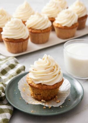 A plate with a carrot cake cupcake with a glass of milk and platter of more cupcakes in the background.