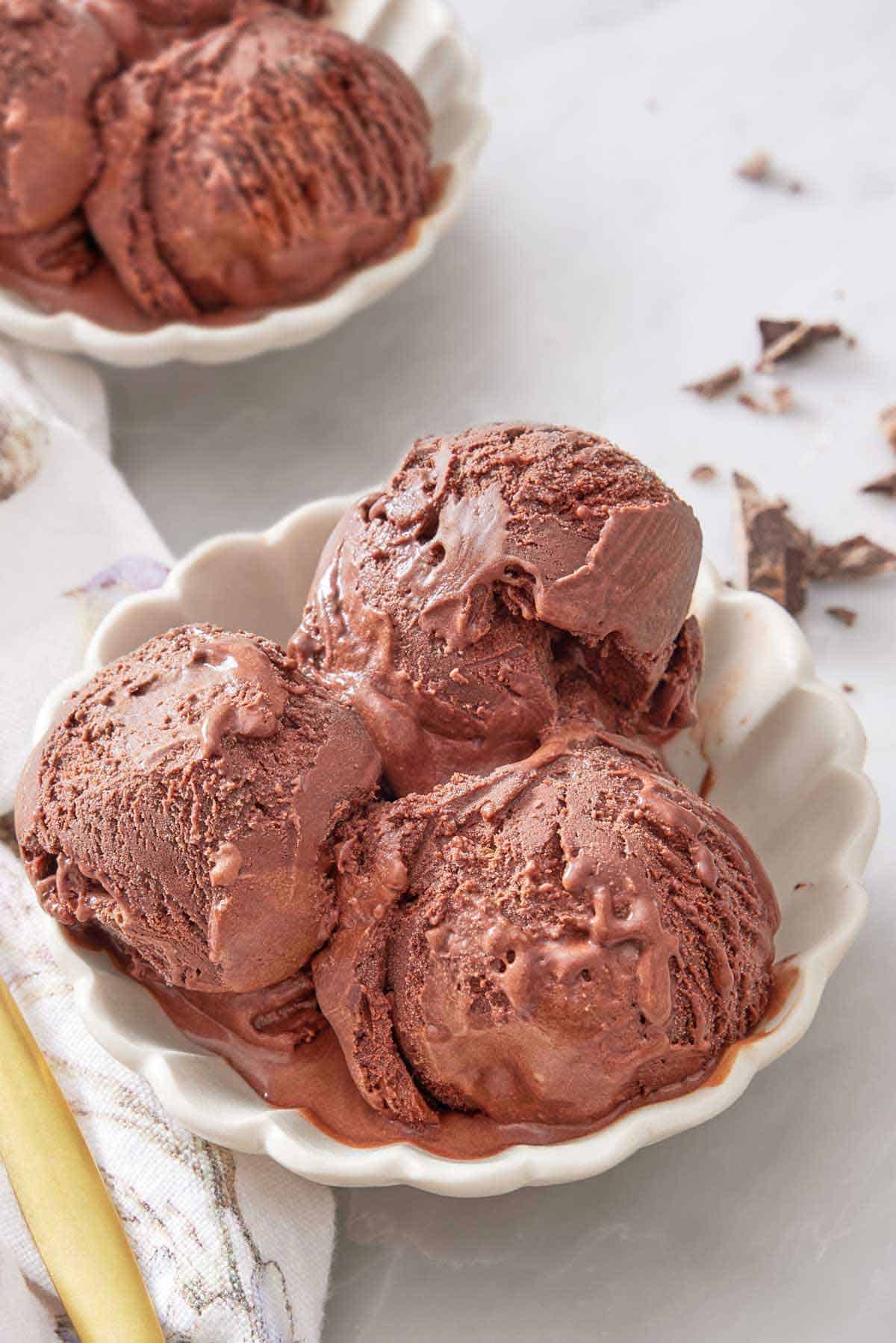A bowl with three scoops of chocolate ice cream with another bowl in the background.