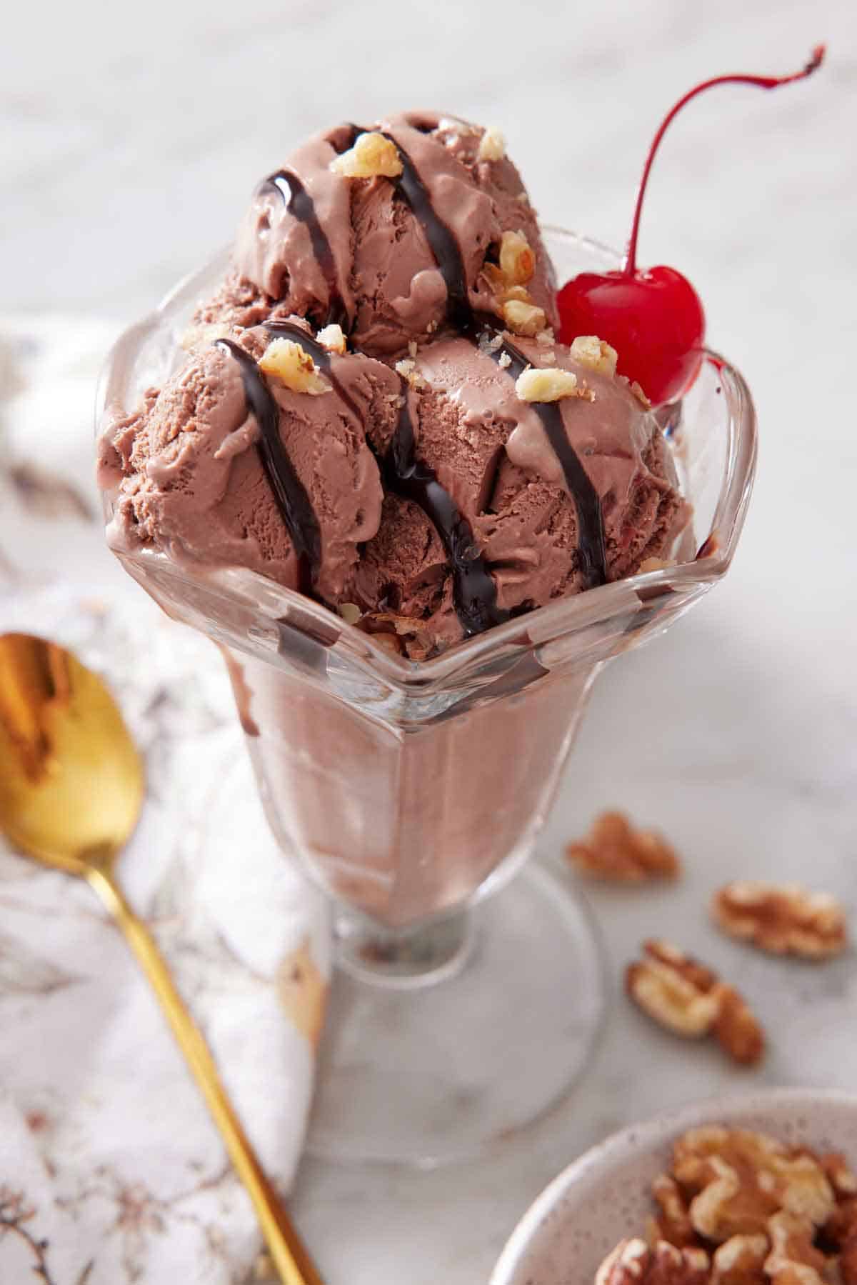A glass with chocolate ice cream topped with a cherry, chocolate drizzle, and crushed nuts.