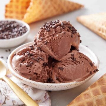 A plate with three scoops of chocolate ice cream topped with chocolate sprinkles. Waffle cones scattered around and a bowl of sprinkles in the back.