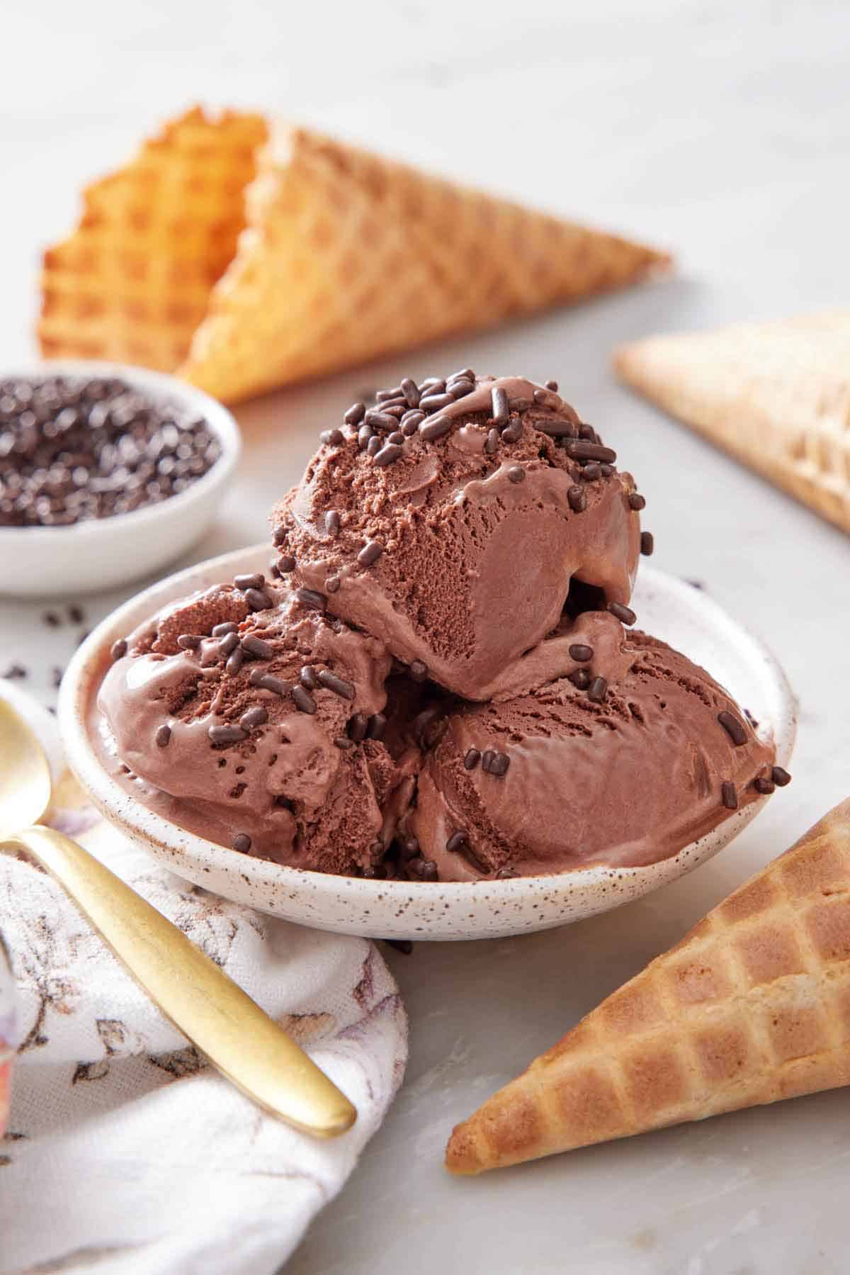 A plate with three scoops of chocolate ice cream topped with chocolate sprinkles. Waffle cones scattered around and a bowl of sprinkles in the back.