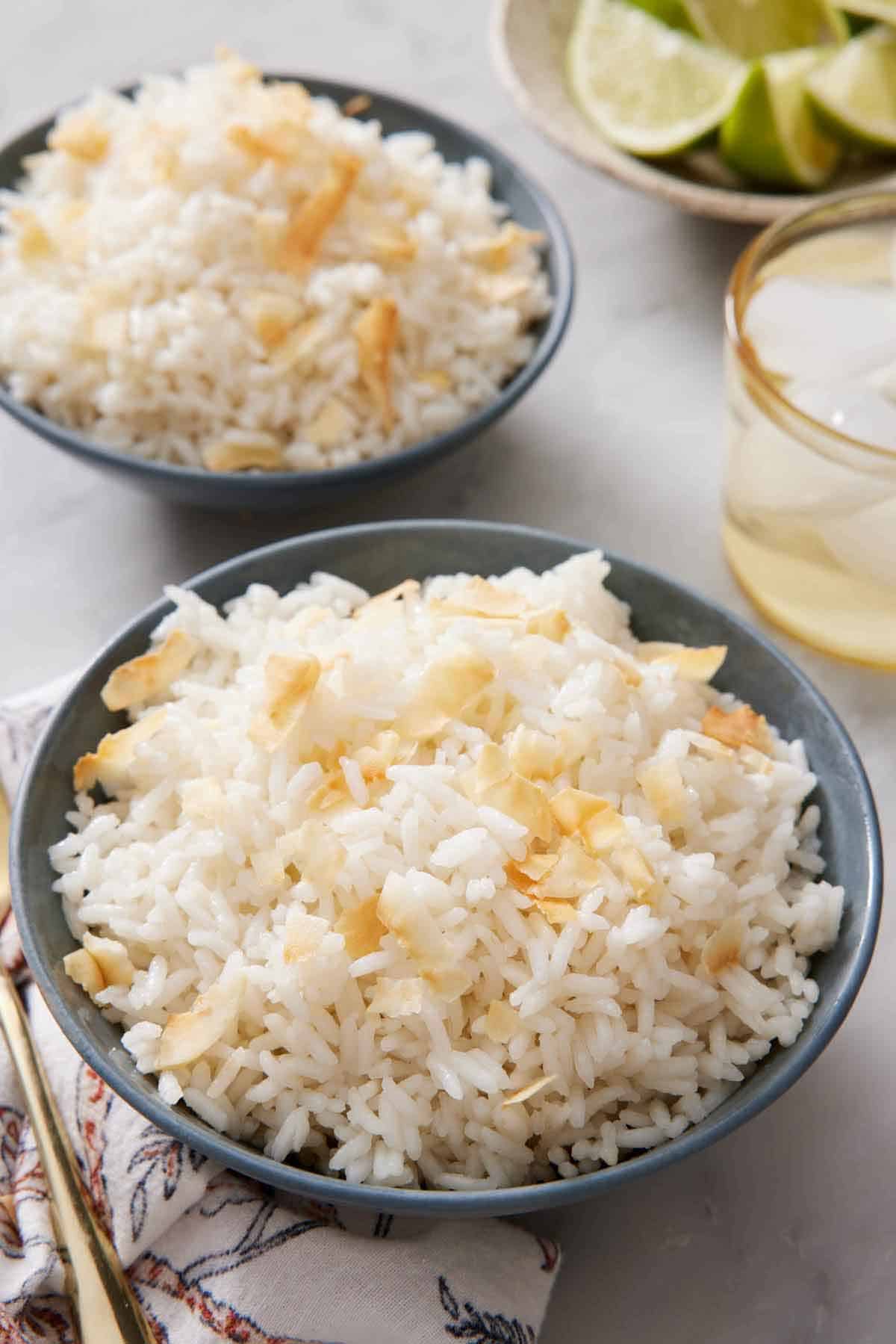 Two bowls of coconut rice topped with toasted coconut. A bowl of lime in the back along with a glass of water.