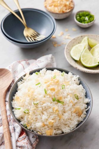 A bowl of coconut rice topped with green onions and toasted coconut. Bowl of limes in the back along with more garnishes and bowls with forks.
